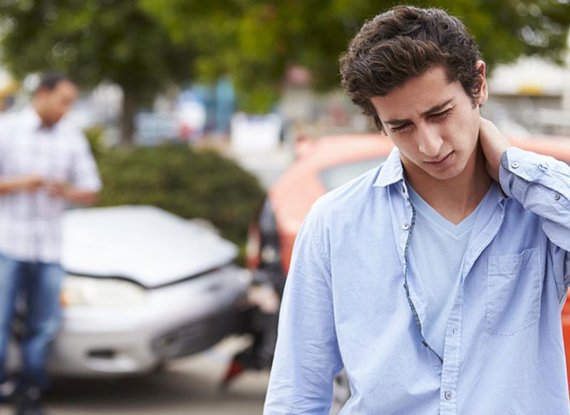 Young man holding his neck in pain in front of a car accident